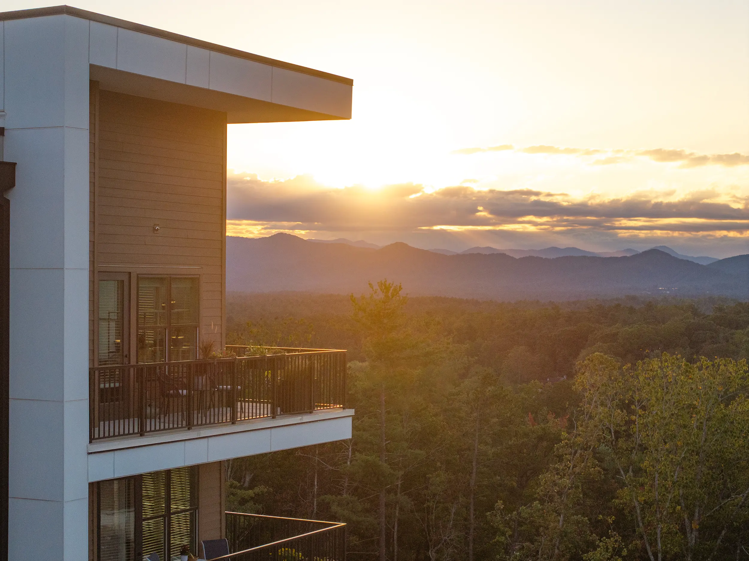 A modern building in the foreground with a balcony overlooking a scenic sunset view. Mountains are visible in the background beneath a colorful sky.