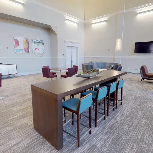 A spacious modern common area with a large table and colorful chairs, seating area, and wall-mounted TV.