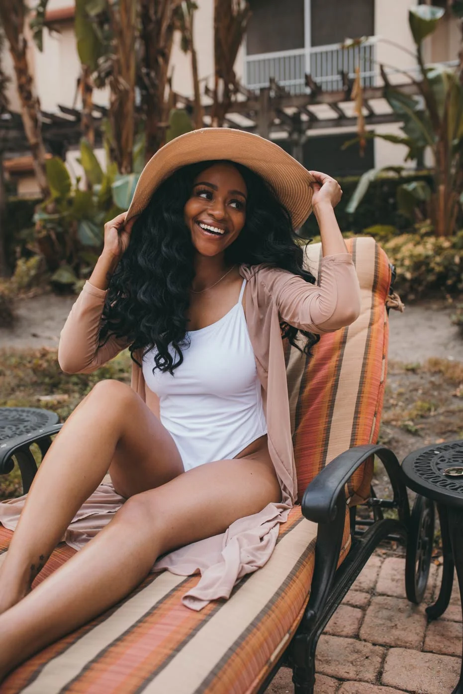 A smiling woman lounging on a chair outdoors, wearing a white swimsuit and a light brown cover-up, complemented by a wide-brimmed hat. The background features tropical plants and a residential area.