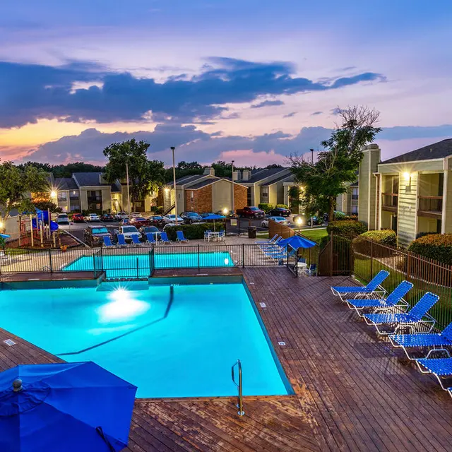A view of a well-lit swimming pool area surrounded by lounge chairs and umbrellas at dusk.