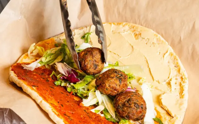 A falafel wrap being assembled with tongs, featuring a flatbread spread with hummus, vegetables like lettuce and cabbage, and three falafel balls.