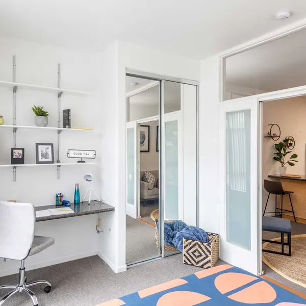 A modern home office with a stylish desk, wall shelving, and an open layout leading to a kitchen area.
