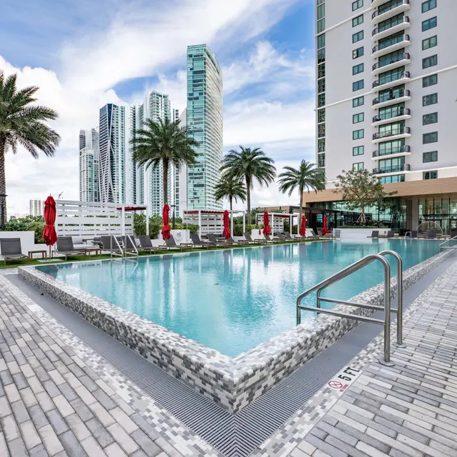 A modern pool area surrounded by palm trees and tall buildings, featuring lounge chairs and umbrellas.