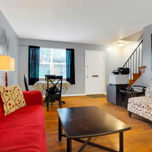 A cozy living room featuring a red sofa with decorative pillows, a dark coffee table in the center, a patterned armchair, and a dining table with chairs near a window. The walls are painted gray and there is a staircase in the background, along with a mirrored wall decor.