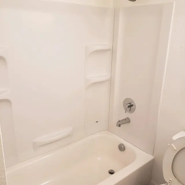 A clean, simple bathroom with a bathtub and shower. The bathtub is beige and features built-in shelves on the wall. A modern showerhead is mounted above. A white toilet is visible beside the bathtub. The walls are painted white.