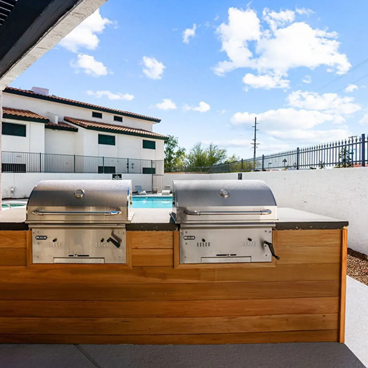 An outdoor grilling area featuring two stainless steel grills situated on a wooden structure. In the background, there is a swimming pool and a residential building visible, under a clear blue sky with scattered clouds.