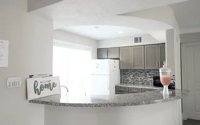 A modern kitchen featuring a curved bar area with a welcome home sign. The kitchen has gray cabinets, a white refrigerator, and a decorative jar filled with a pink drink. Natural light floods in from a window nearby, highlighting the sleek design.