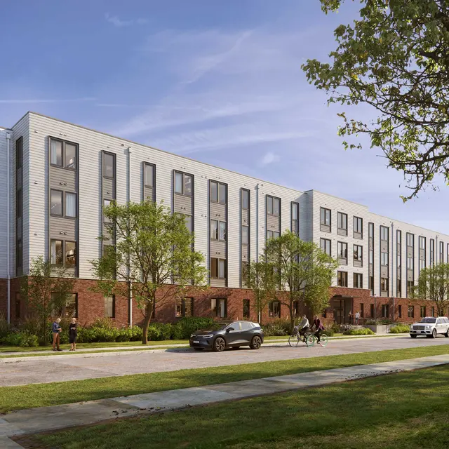 A modern, multi-story building with a combination of brick and colored panel exterior, surrounded by greenery and cars parked nearby. People are walking on the path in front of the building.