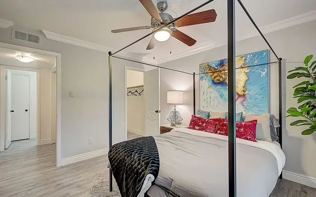 A modern bedroom featuring a four-poster bed with colorful pillows, a ceiling fan, a bright artwork above the bed, and a potted plant. Soft gray walls and light wood flooring enhance the cozy atmosphere.