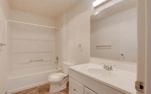 A clean and simple bathroom featuring a white bathtub, toilet, and a large mirror above the sink. The walls are painted a light color, and there is a towel rack on the wall.