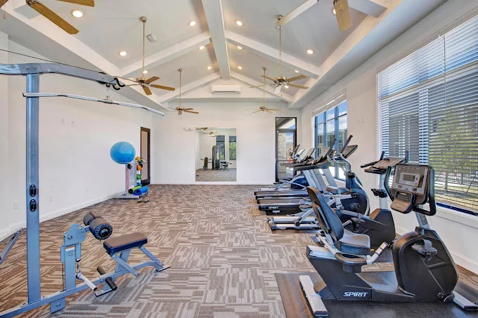 A spacious indoor gym featuring exercise equipment, large windows, and ceiling fans.
