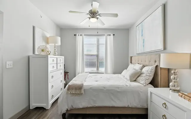 Cozy bedroom with a beige bed, white dresser, lamps, a ceiling fan, and light curtains letting in natural light.