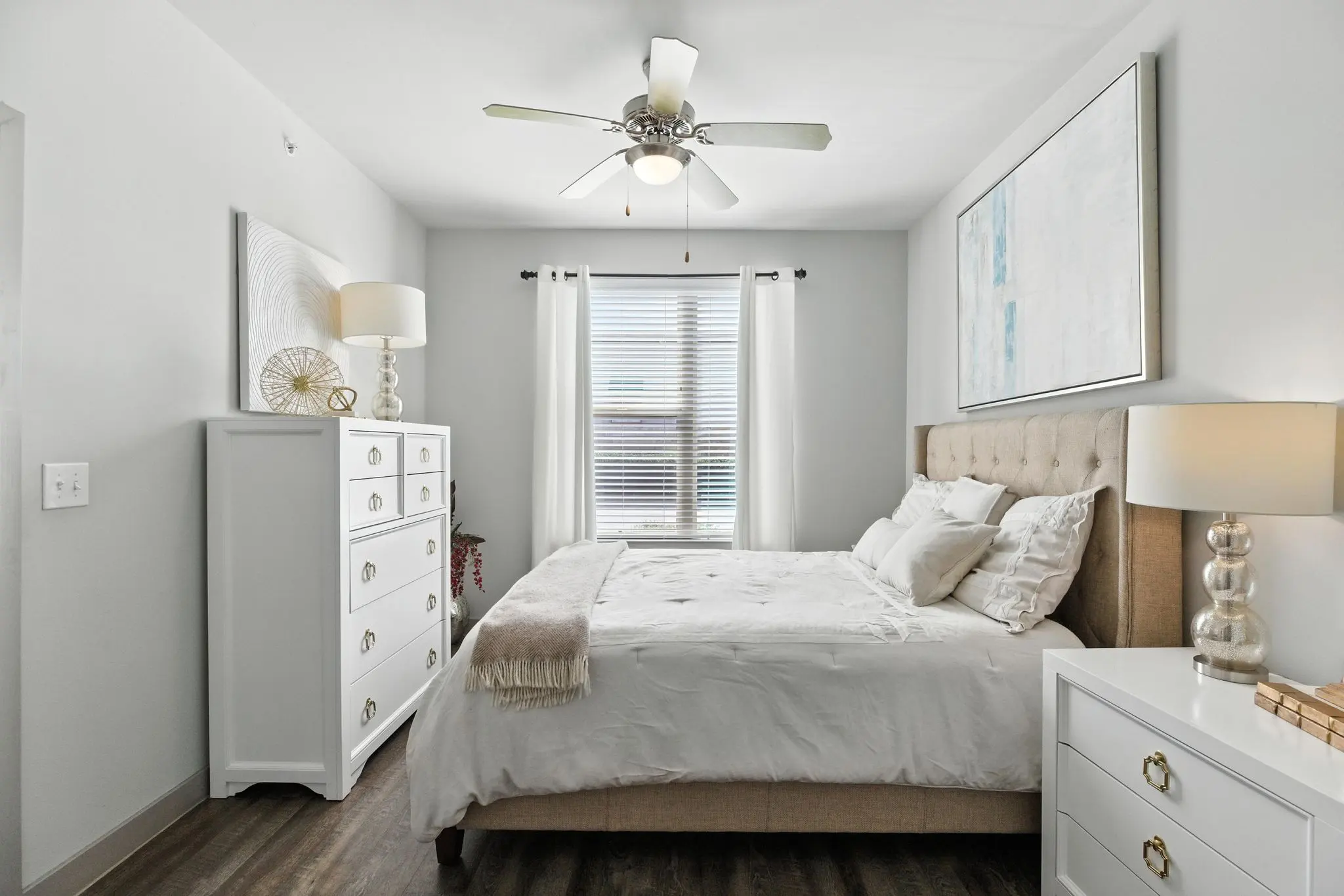 Cozy bedroom with a beige bed, white dresser, lamps, a ceiling fan, and light curtains letting in natural light.