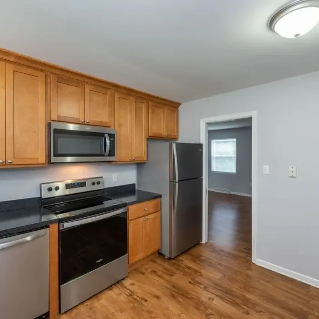 A modern kitchen featuring wooden cabinets, stainless steel appliances, and a black countertop. The room has a light-colored wall and hardwood floor, with a doorway leading to another room.
