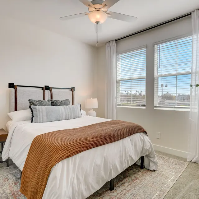 Cozy Modern Bedroom A cozy modern bedroom featuring a comfortable bed with white linens and an orange throw blanket, light wooden furniture, and large windows with sheer curtains.