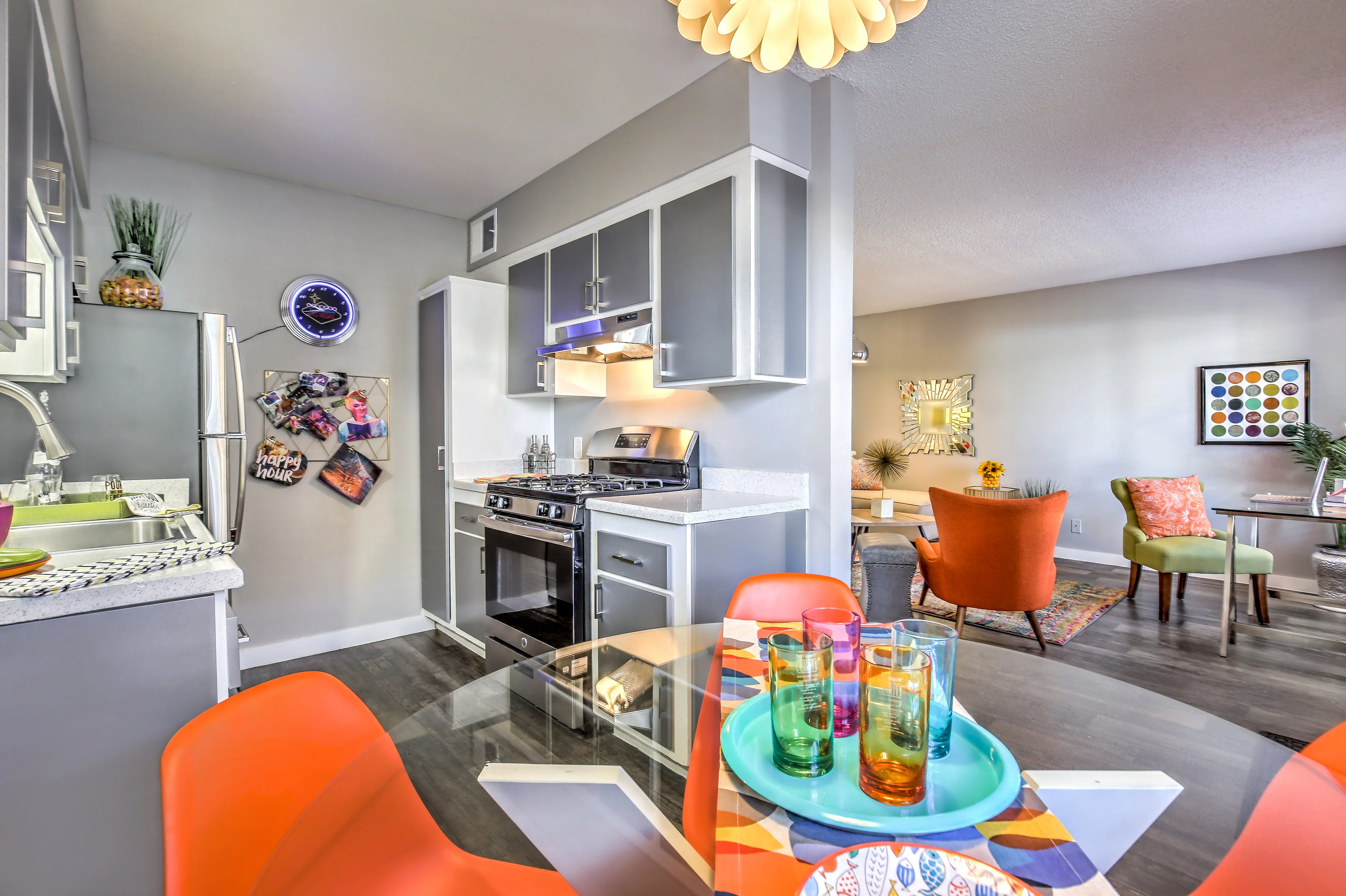 A modern kitchen area with colorful decor, featuring orange chairs and a glass dining table with vibrant glasses on it. The kitchen is equipped with stainless steel appliances and has a light-filled living space beyond.