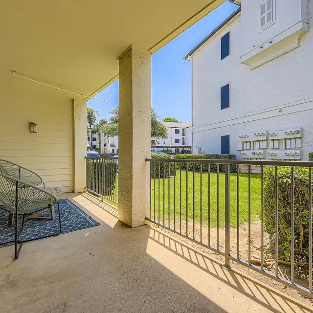 A view from an apartment balcony featuring a patterned rug, two green chairs, and a railing overlooking a green lawn and another building in the background.