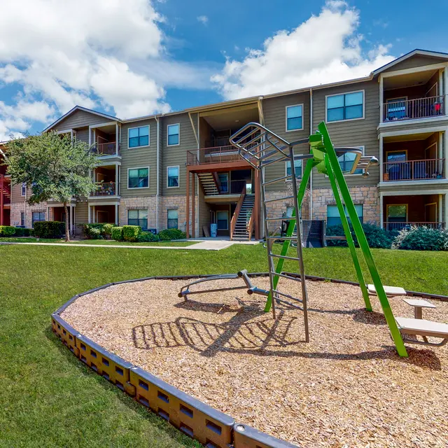Playground Near Apartment Complex A playground area with a swing set and a landscaped apartment complex in the background under a blue sky with clouds.
