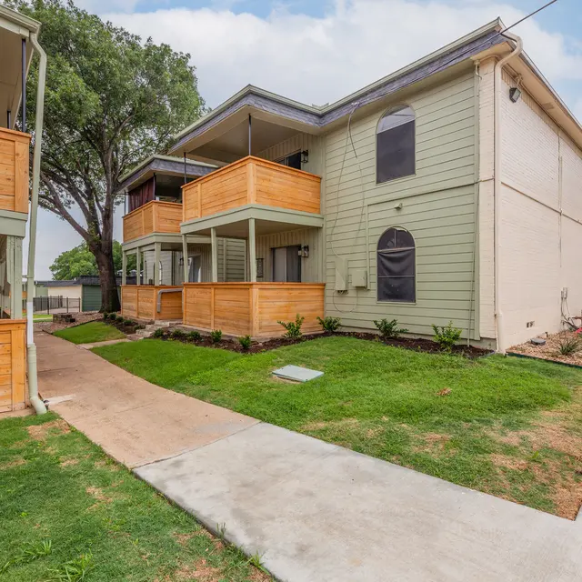 The Hills - Exterior view of a multi-unit apartment building with wooden decks and green siding, surrounded by grass and trees.