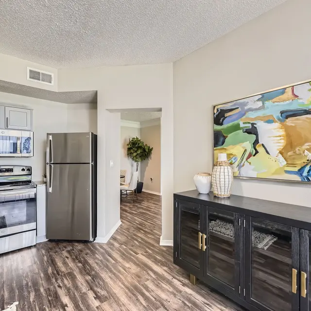 A modern kitchen with stainless steel appliances, a black wooden cabinet, and colorful wall art on a beige wall. The floor features dark wooden planks, and there's a plant visible in the background.