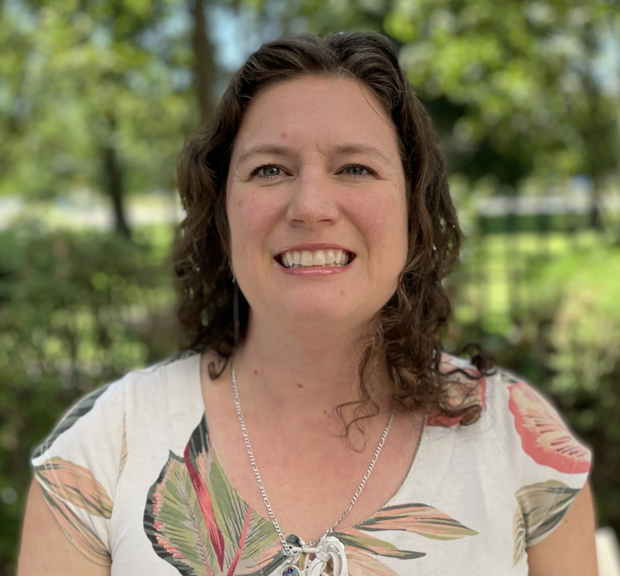 Outdoor Smile A woman with curly hair smiling outdoors, wearing a floral-patterned shirt. Green trees and foliage are in the background.