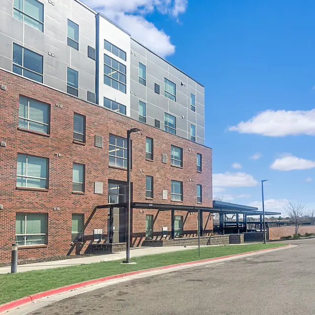 A modern building with a mix of brick and gray paneling, featuring multiple windows. A sidewalk runs alongside the building and a small lawn is in front. The sky is bright with a few clouds.