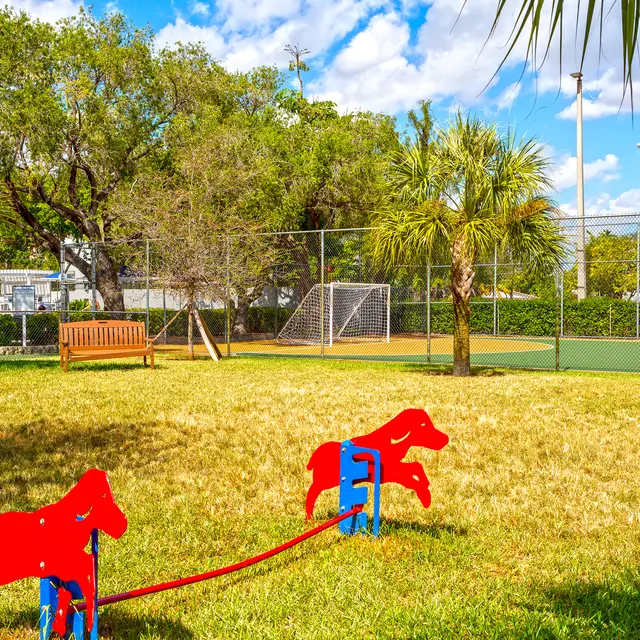 Dog Park 1 - Biscayne Apartments A dog park featuring red dog-shaped play equipment, a bench, and green grass under a clear blue sky with a few clouds.