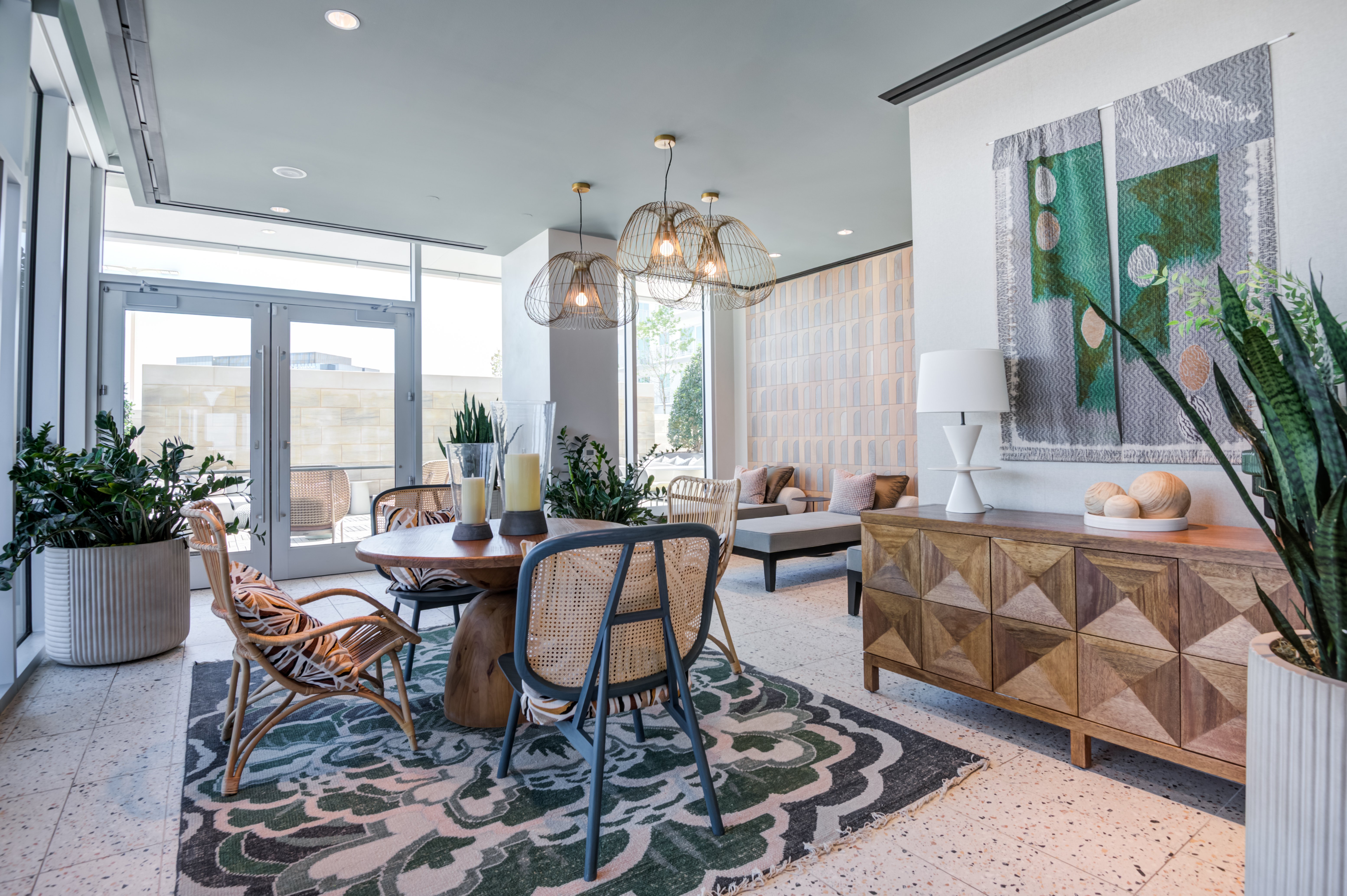 Modern Living Space with Natural Light A stylish modern living space featuring a wooden dining table surrounded by woven chairs, a patterned area rug, and potted plants. Large windows allow natural light in, with decorative wall art and a wooden sideboard adding elegance.