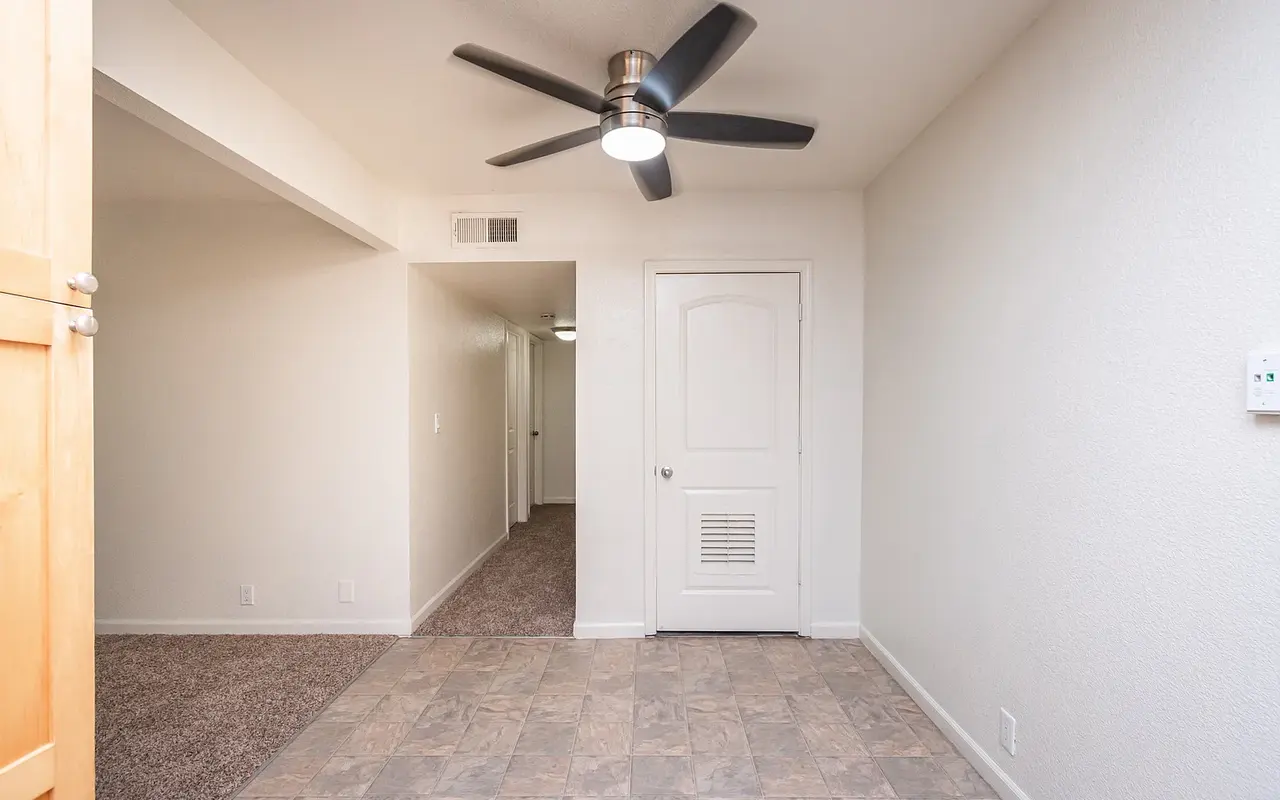 A view of an apartment entryway featuring a tiled floor and a ceiling fan. The walls are painted white, and there is a door on the right leading to another room. A hallway is visible in the background.