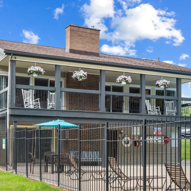 A two-story community building with a porch featuring rocking chairs and flower baskets, surrounded by a fence. Below the porch, there's a patio area with lounge chairs and an umbrella.