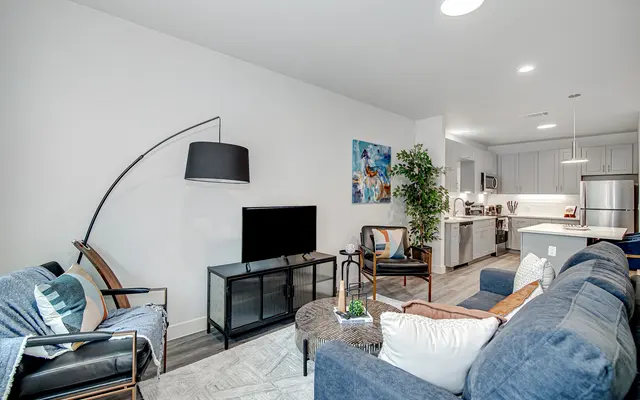 A modern living room featuring a stylish blue sofa, an elegant armchair, a black floor lamp, and a flat-screen TV on a stand. The kitchen is visible in the background with modern appliances. There's a potted plant adding greenery to the space.