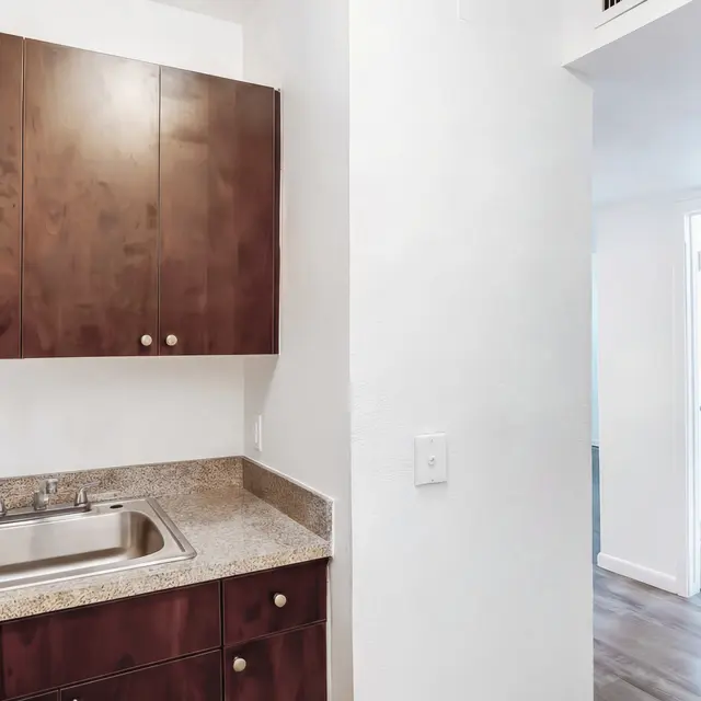 A small kitchen area featuring dark wood cabinets above a granite countertop with a sink. A doorway leads to another room in the background.