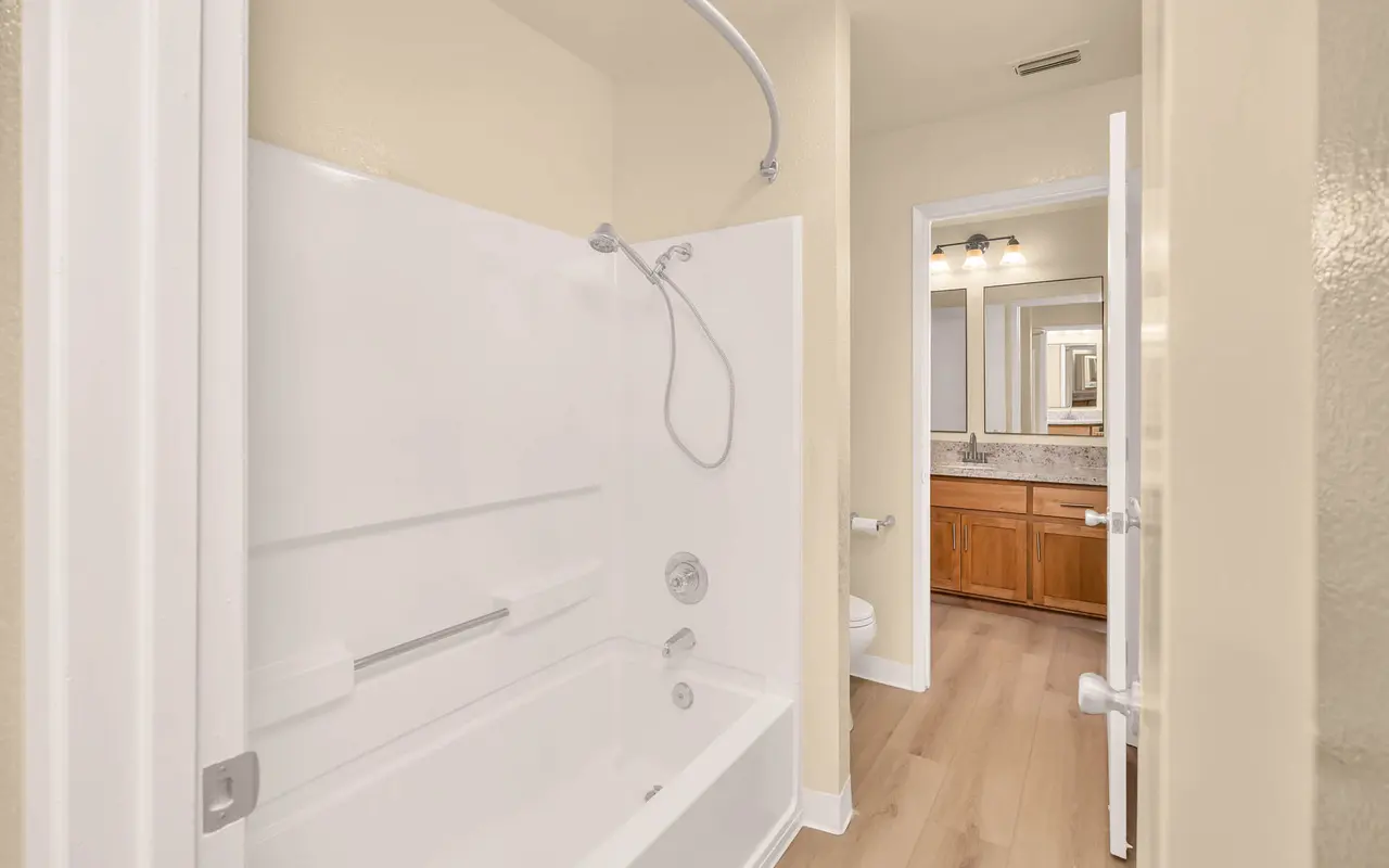 Interior view of a modern bathroom with a bathtub and shower combination, wooden flooring, and a mirror visible in the background.