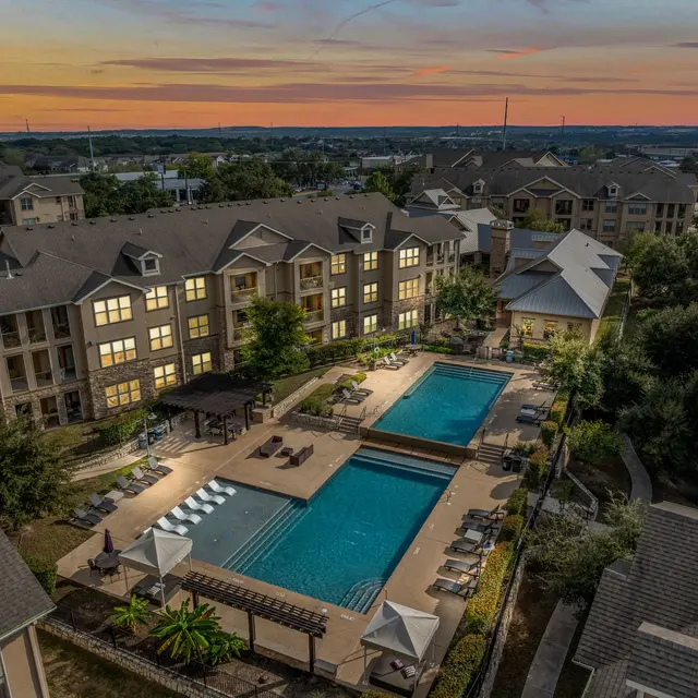 Aerial view of an apartment complex at sunset featuring a swimming pool area surrounded by lush greenery and lounge chairs.