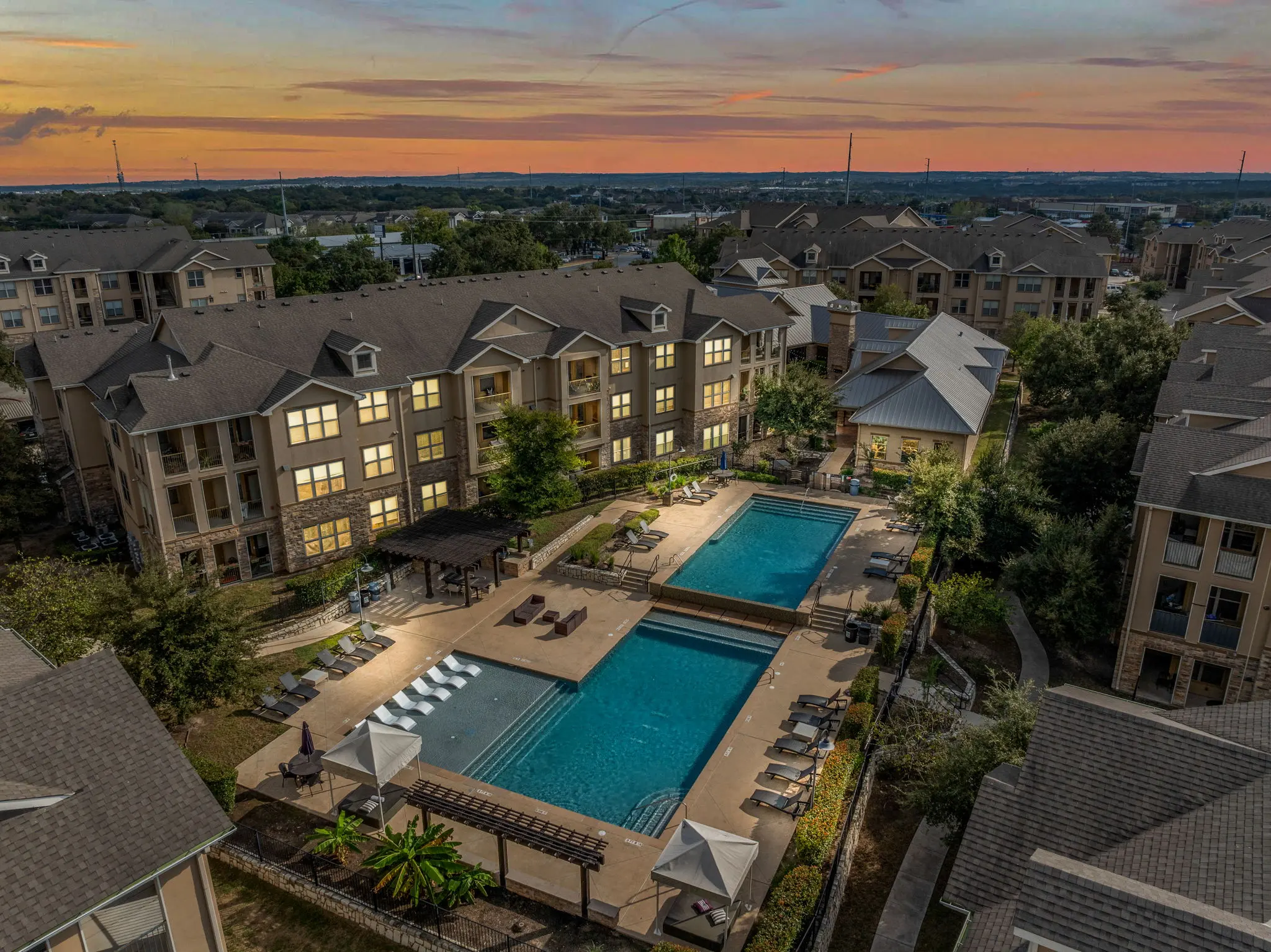 Aerial view of an apartment complex at sunset featuring a swimming pool area surrounded by lush greenery and lounge chairs.