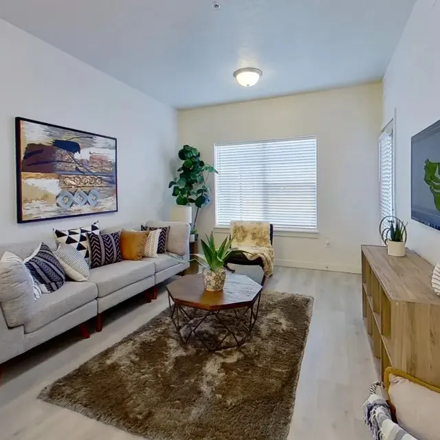 A cozy, modern living room featuring a light-colored couch with decorative pillows, a coffee table with a plant, a rug, and a wooden sideboard with decor items. Large windows allow natural light to enter the space.