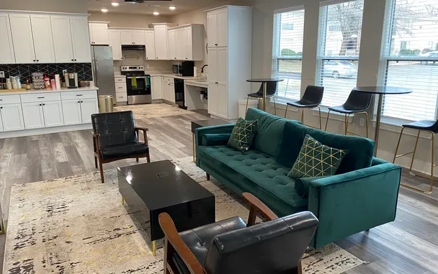A modern living room featuring a teal couch, two black chairs, and a black coffee table on a patterned rug. The kitchen area is in the background with white cabinets and appliances. Large windows provide natural light.