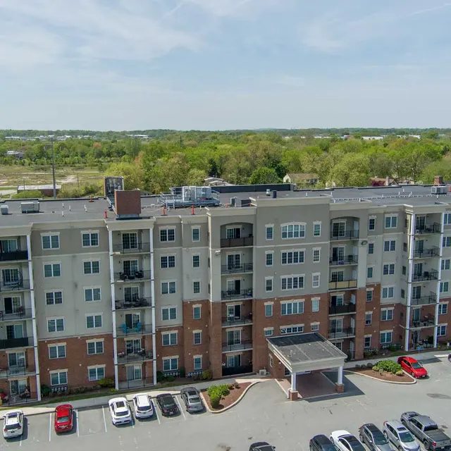 Aerial view of a modern apartment complex featuring five stories with a mix of brick and siding, surrounded by a parking lot with several vehicles.