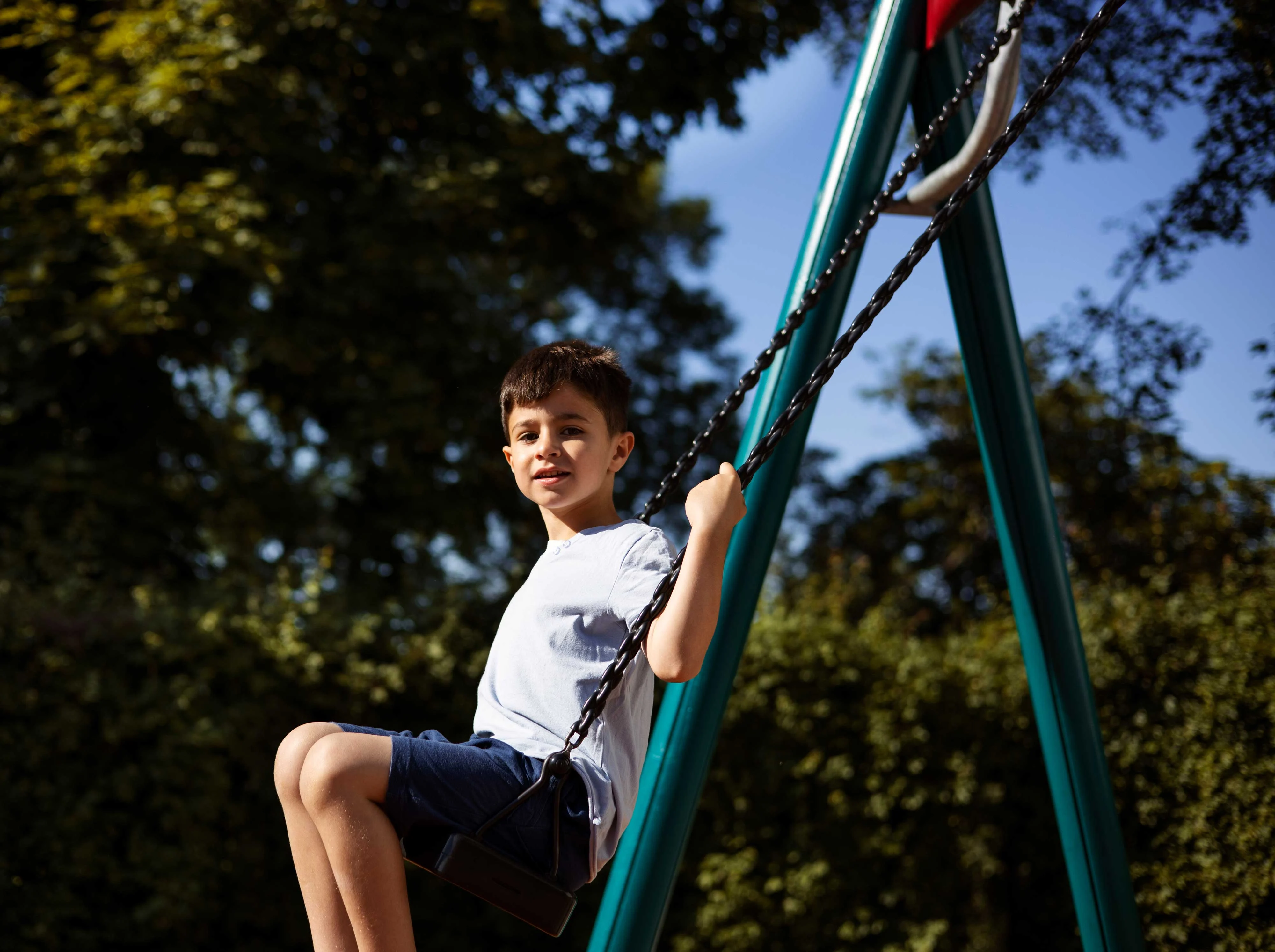 A young boy sitting on a swing in a sunny park, smiling at the camera with trees in the background.