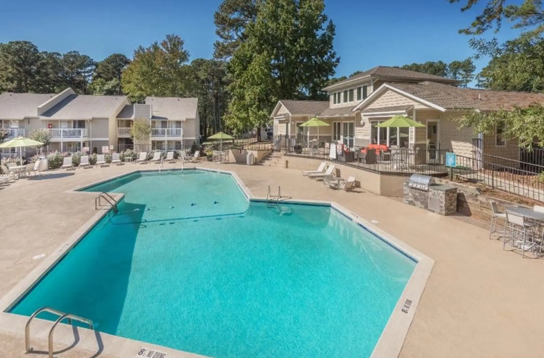 A bright and inviting outdoor pool area surrounded by lounge chairs, with a shaded seating area and lush trees in the background.