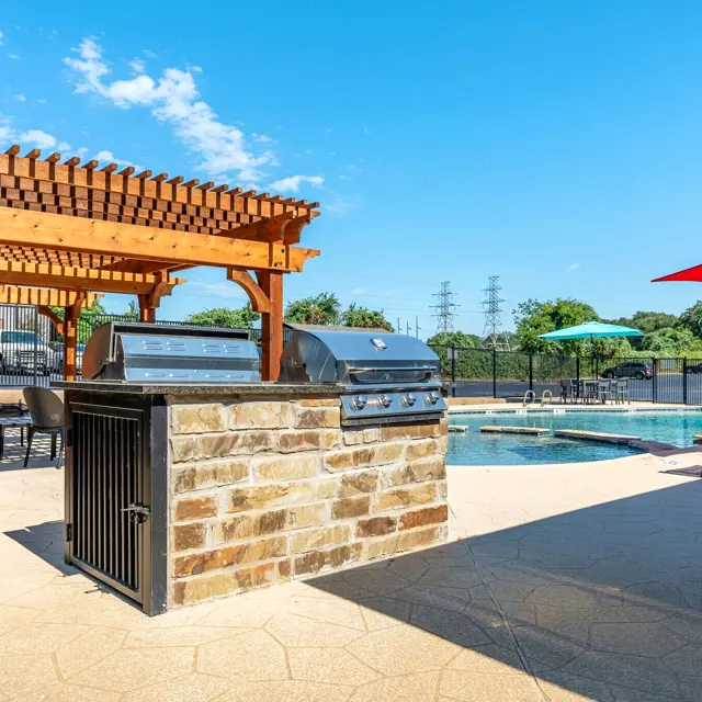 Outdoor pool area featuring a stone grill, wooden pergola, lounge chairs, and umbrellas