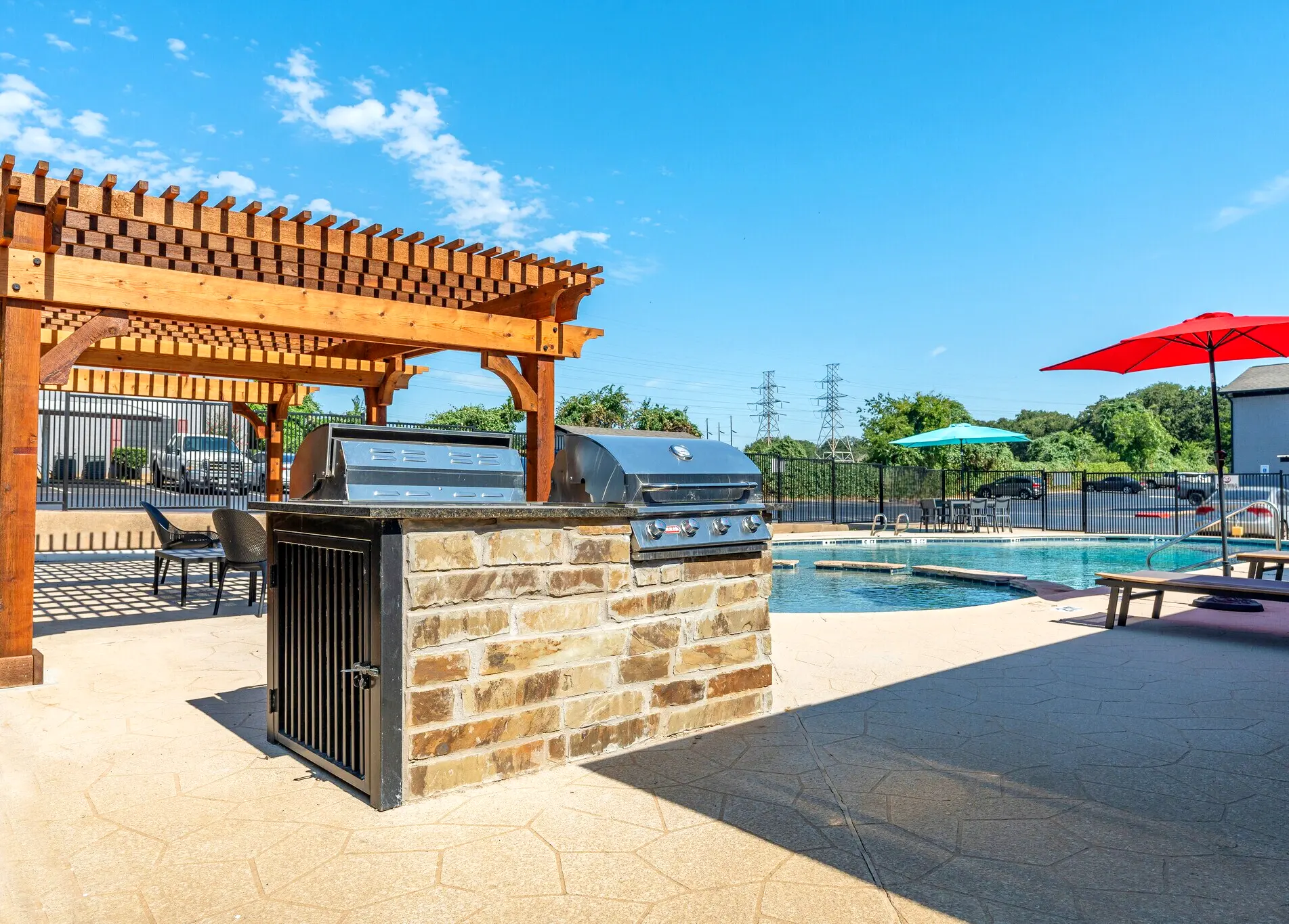 Outdoor pool area featuring a stone grill, wooden pergola, lounge chairs, and umbrellas