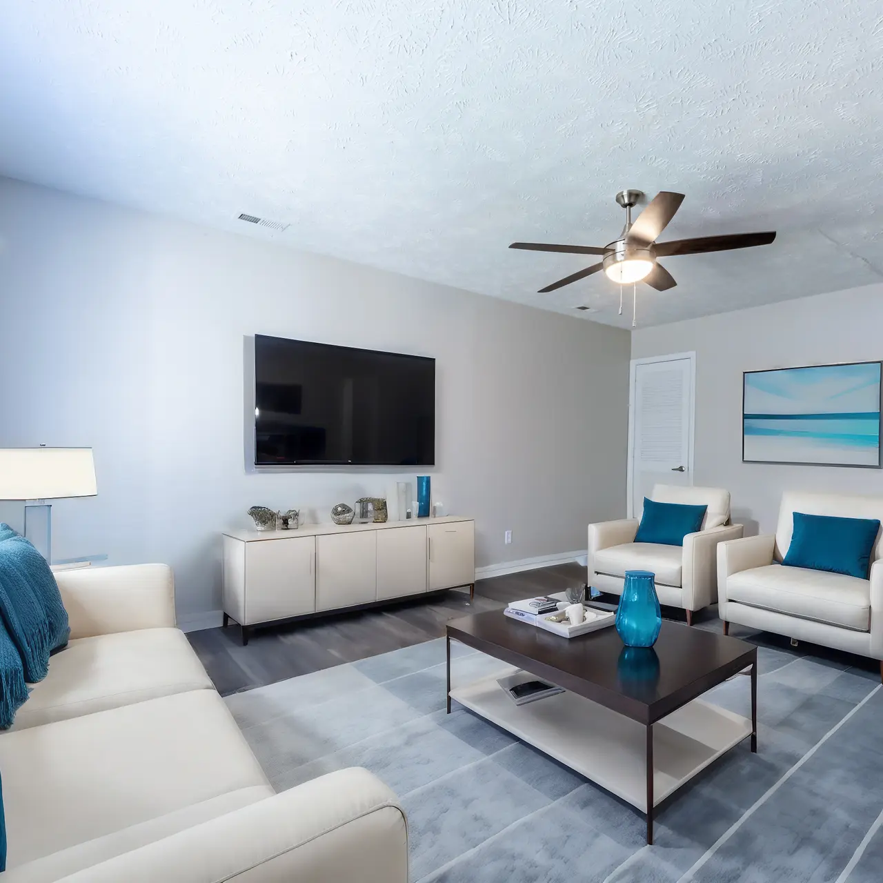A modern living room featuring a light color scheme with a beige couch and two light-colored armchairs. A coffee table sits in the center of the room with decorative items. There is a large flat-screen TV mounted on the wall, and large windows provide natural light.