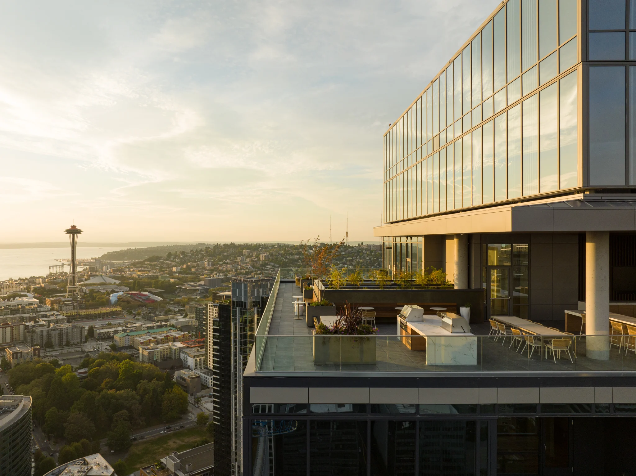 A panoramic view from a rooftop terrace showcasing a city skyline with a prominent landmark, with the sea and distant mountains in the background during sunset.