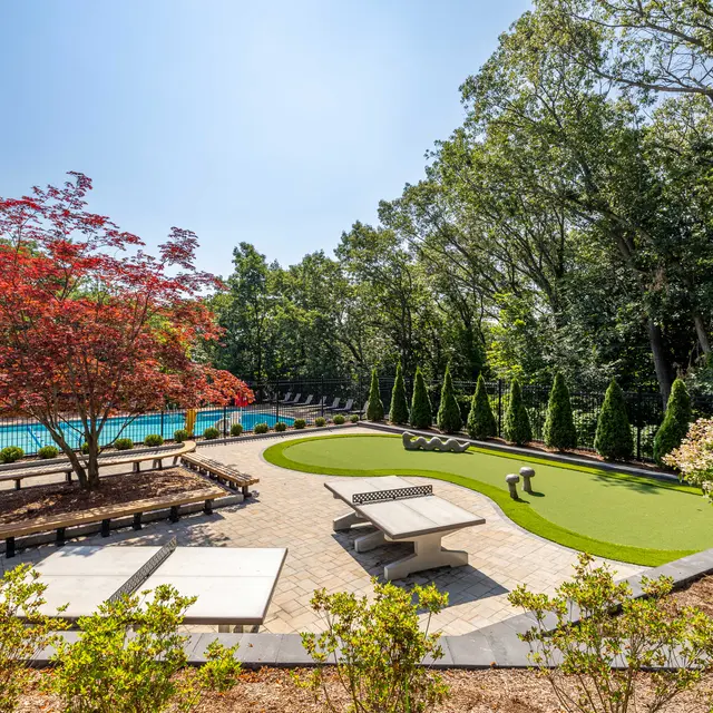 A serene outdoor space featuring a swimming pool, greenery, and a vibrant red tree. The area includes seating, a small green lawn, and a stone table.