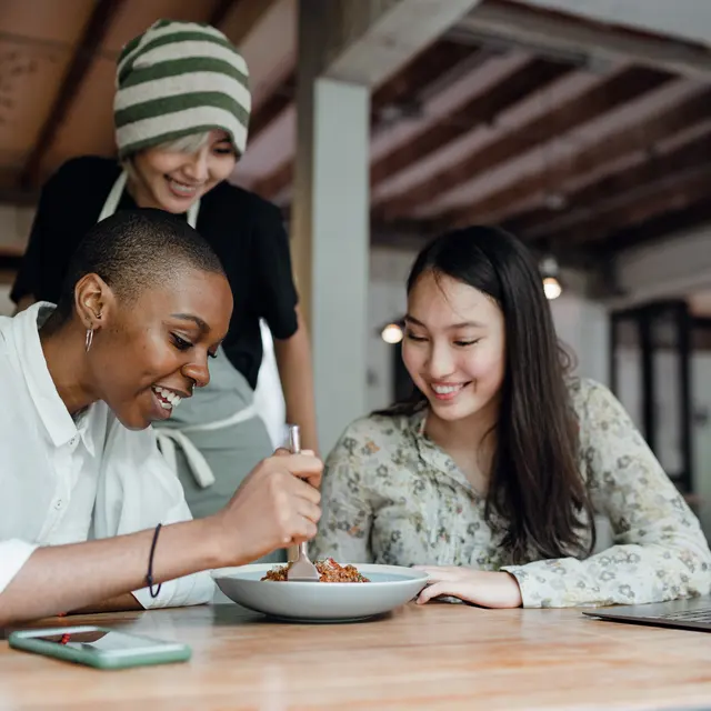 Three people in a cozy café, with one woman about to eat from a bowl, while two others watch with smiles. A laptop sits on the table.