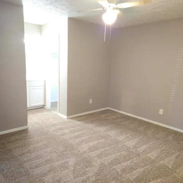 A spacious living area featuring light beige walls, a ceiling fan, and new carpet. A doorway leads to a well-lit area with a sink and cabinets.