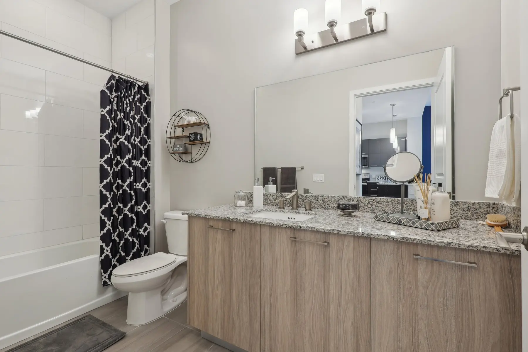 A modern bathroom featuring a wooden vanity with a granite countertop, a large mirror above, and a spacious shower area with a white bathtub. The bathroom has a black and white patterned shower curtain, neatly arranged toiletries, and a contemporary light fixture.