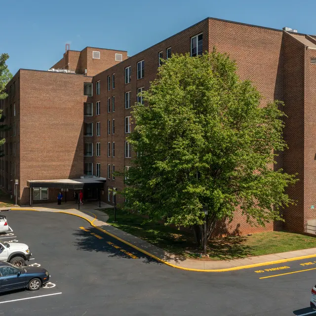 A five-story brick apartment building surrounded by trees and a parking lot. Several cars are parked in front, with green foliage nearby and a clear blue sky overhead.