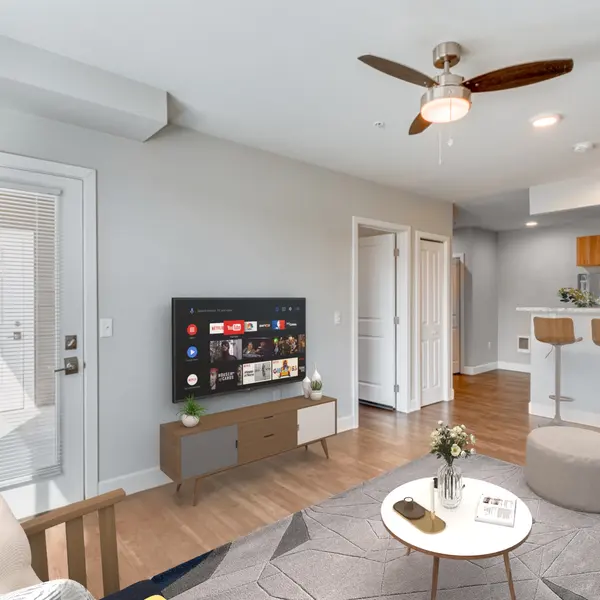 A bright, open-concept living room featuring a mounted smart TV, a cozy seating area, and a view into a modern kitchen with bar seating. The space includes wood flooring, a ceiling fan, and a back door with natural light streaming in.